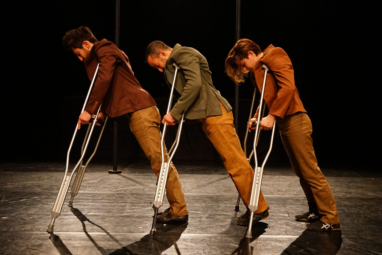 Trois interprètes en veste et pantalon de velours, penchés en avant sur des béquilles, sur un plateau noir.